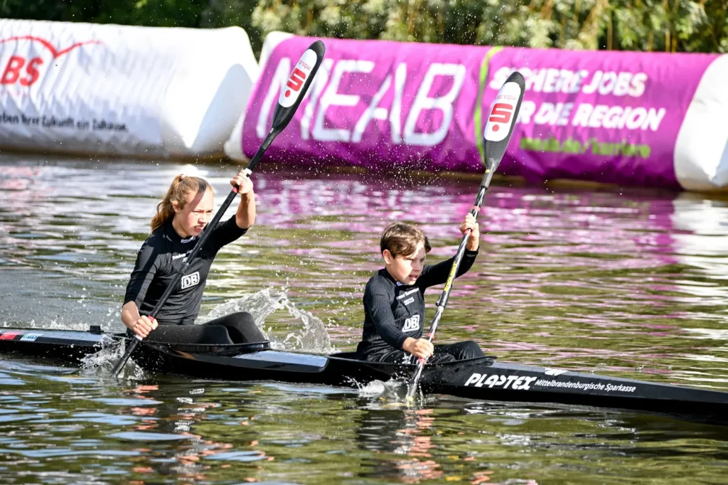 Zwei Nachwuchssportlerinnen im Zweier-Kajak vor den Sponsorenbahnen des Kanusprints