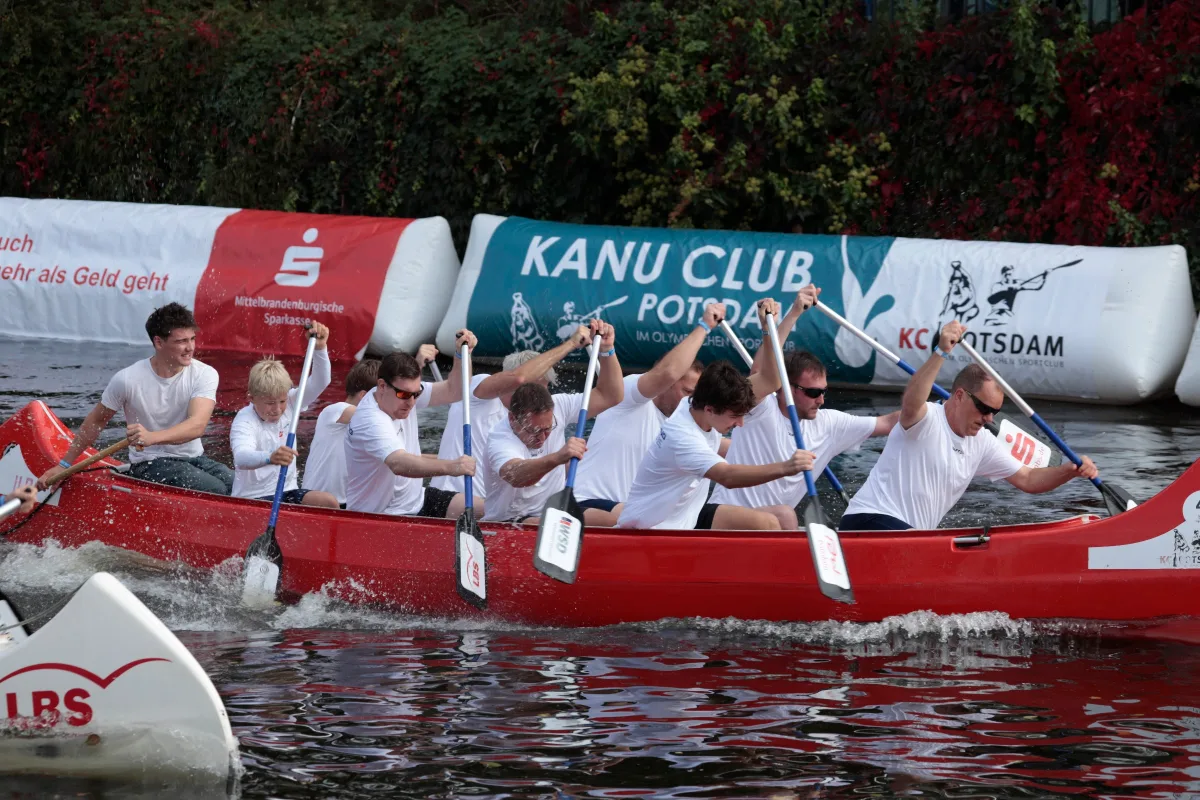 Sponsorenrennen: Team in weißen Shirts im roten Drachenboot vor Banden des Kanu Club Potsdam